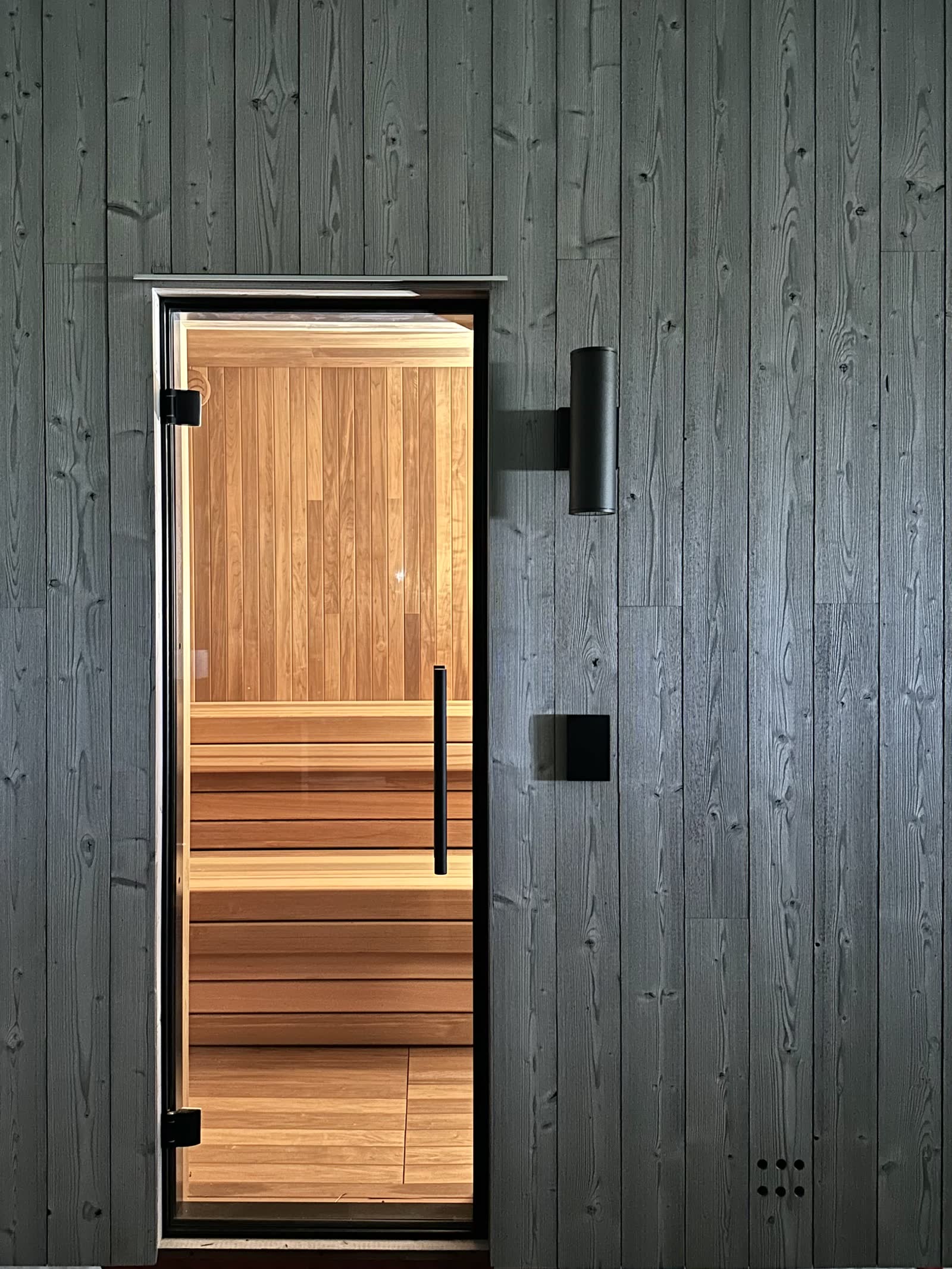 Outdoor sauna with dark wood cladding and a glass door, warm-lit wood benches inside